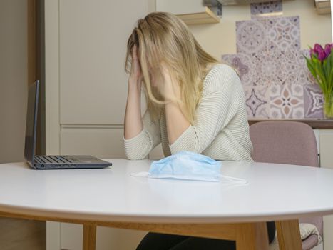 Beautiful young woman working on laptop computer while sitting at living room At Home. Medical blue mask on the table. Depressed And Sad girl In Kitchen. Health care and antivirus protection concept