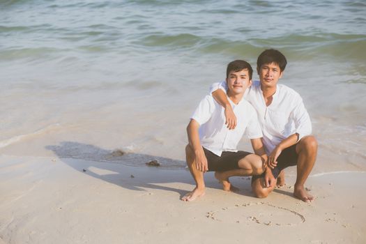 Young asian couple gay smiling romantic drawing heart shape together on sand in vacation, homosexual happy and fun hug with love sitting on sand at the beach in travel summer, LGBT legal concept.
