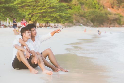 Homosexual portrait young asian couple sitting hug and pointing something together on beach in summer, asia gay going tourism for leisure and relax with happy in vacation at sea, LGBT legal concept.