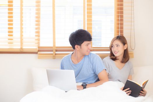 Asian young couple with man using laptop computer and searching internet and woman reading notebook on bed at bedroom with happy and relax, communication and business concept.