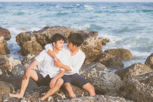 Homosexual portrait young asian couple sitting hug together on rock or stone in the beach in summer, asia gay going tourism for leisure and relax with happy in vacation at sea, LGBT legal concept.