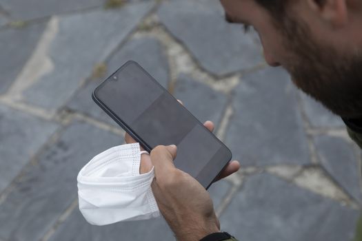 A young man, grabbing a protective face mask, holds a smart mobile phone with his hand, Spain.