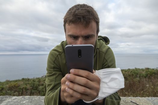 A young man, grabbing a protective face mask, holds a smart mobile phone, with the sea behind him, Spain.