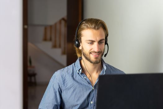 Blond male working from home with headphones using wireless internet on laptop, man call center agent or telemarketer