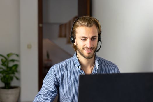 Blond male working from home with headphones using wireless internet on laptop, man call center agent or telemarketer