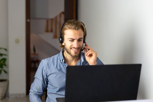 Blond male working from home with headphones using wireless internet on laptop, man call center agent or telemarketer