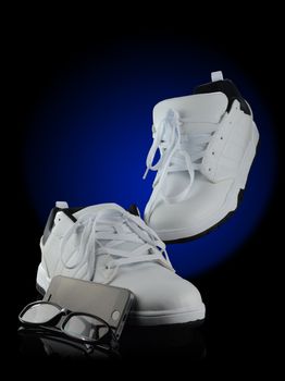 A poster style image of a pair of mens white street styled shoes with a cell phone and eye glasses over a black reflective background with a blue spot color for added depth.
