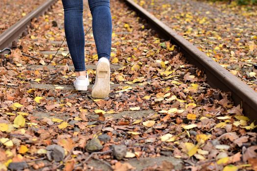 Walking legs wearing blue jeans and white shoes on autumn leaves and old railroad tracks