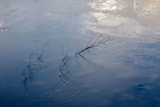 The surface of a pond is freezing over as fall turns to winter. Ice is forming across the water, with fallen tree branches emerging from it.
