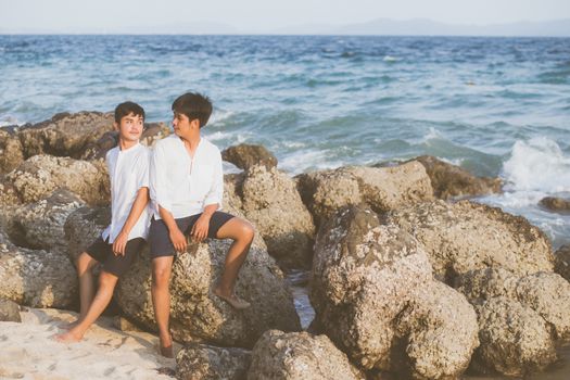 Homosexual portrait young asian couple sitting hug together on rock or stone in the beach in summer, asia gay going tourism for leisure and relax with happy in vacation at sea, LGBT legal concept.