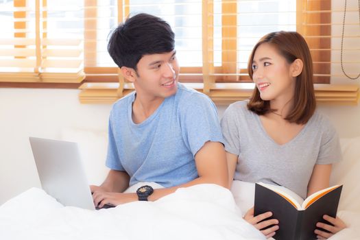 Asian young couple with man using laptop computer and searching internet and woman reading notebook on bed at bedroom with happy and relax, communication and business concept.