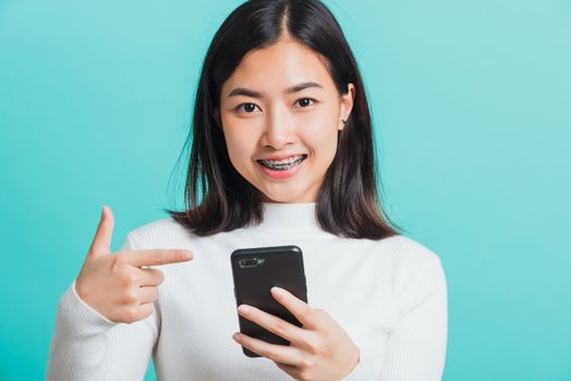 Beautiful Asian woman smile she pointing finger to a smartphone, female excited cheerful after received promotion her point finger on mobile phone isolated on a blue background, Technology concept