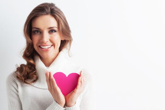 Pretty woman in white winter sweater holding pink paper heart symbol of Valentine day over white background