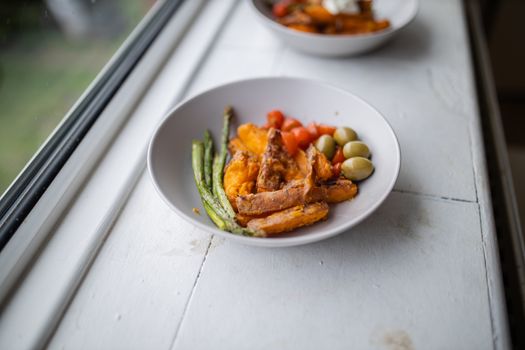 Sweet potato fries with cottage cheese on top, olives, tomatoes, and asparagus dish next to window. Nutritious dish with vegetables on white plate. Balanced diet