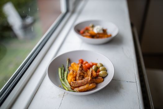 Sweet potato fries with cottage cheese on top, olives, tomatoes, and asparagus dish next to window. Nutritious dish with vegetables on white plate. Balanced diet