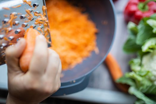 Female hands firmly grating carrot on metal grater and into bowl. Shredded carrots in bowl surrounded by vegetables. Vegan meal preparation