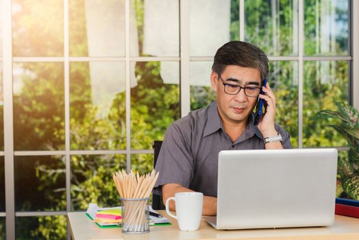 Asian executive senior businessman sitting on desk office he using his mobile phone and talking with somebody, the confident middle aged handsome man using laptop computer at workplace home office