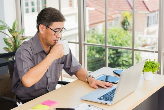 Asian executive senior businessman sitting on desk office with his office, the confident middle aged handsome man using laptop computer at workplace home office and drinking a cup of coffee