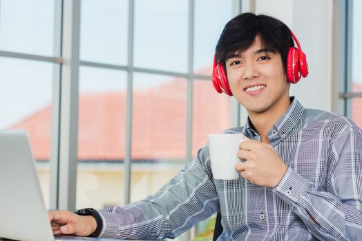 Asian young businessman happiness with red headphones sitting on desk workplace home office with a laptop computer, confident handsome man lifestyle smile relax holding a coffee cup, listens to music