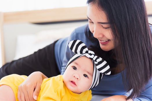 Portrait of beautiful young Asian mother playing and smiling together with his newborn little baby at home, Parent mom and little kid relaxing in the bedroom, Family having fun together