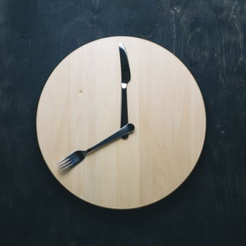 Intermittent fasting and skip breakfast concept - empty wooden round tray or trencher with cutlery as clock hands on dark background. Eight hour feeding window concept or breakfast time concept