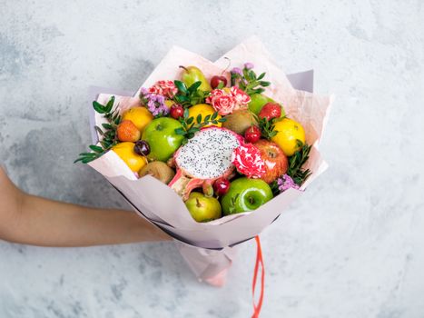 Fruit and berries bouquet. Eating bouquet in female hand on gray background. Apple, orange, strawberry, pear, kiwi, dragon fruit and flowers, eucalyptus. Shallow DOF. Copy space