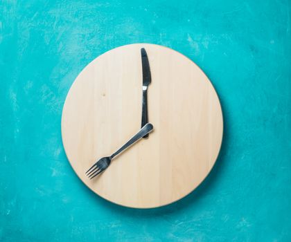 Intermittent fasting and skip breakfast concept - empty wooden round tray or trencher with cutlery as clock hands on blue background. Eight hour feeding window concept or breakfast time concept