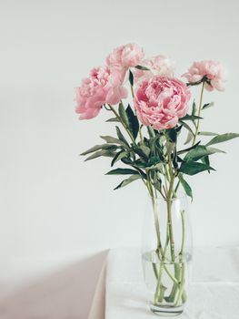 Beautiful pale pink peony bouquet in glass vase on table with white linen tablecloth. Vertical. Copy space for text.