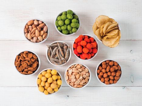 Assortment of different snack for beer, wine, party. Peanuts in coconut glaze, green vasabi, red spicy chilli, yellow cheese glaze, chips, pistachio, crackers, fish on white wooden table. Copy space