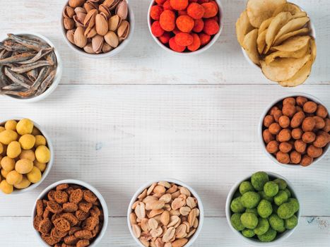 Assortment of different snack for beer, wine, party. Peanuts in coconut glaze, green vasabi, red spicy chilli, yellow cheese glaze, chips, pistachio, crackers, fish on white wooden table. Copy space