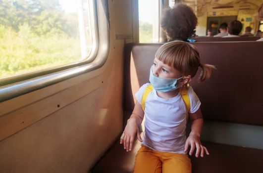 Little girl in a white T-shirt and jeans and a medical mask is sitting in the train. Keep social distancing to avoid the spread of COVID-19. Safe travel.