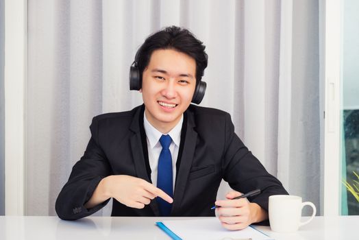 Work from home, Asian young businessman wear suit video conference call or facetime he smiling sitting on desk wearing headphones and raise his hand point finger to a paper job he looking to camera