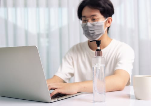 Asian business young handsome man glasses, wearing protective mask with disinfectant gel beside he work from home office remote online with laptop computer quarantines disease coronavirus or COVID-19