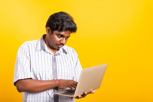 Asian happy portrait young black man smiling standing wear shirt holding and typing laptop computer he looking to camera isolated, studio shot yellow background with copy space