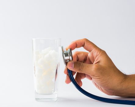 Hand of doctor hold stethoscope check on glass full of white sugar cube sweet food ingredient, isolated on white background, health high blood risk of diabetes and calorie intake unhealthy drink