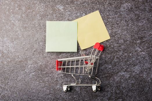 Above blank paper stick note list yellow and green with the shopping cart on concrete background, Shopping reminder concept