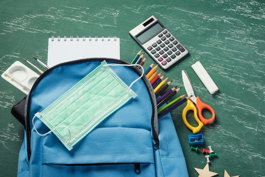 Top view of stylish blue school bag backpack at a green chalkboard with face mask protection and stationery, Back to school education new normal during outbreak COVID-19 or coronavirus concept