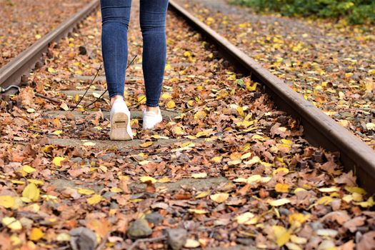 Walking legs wearing blue jeans and white shoes on autumn leaves and old railroad tracks