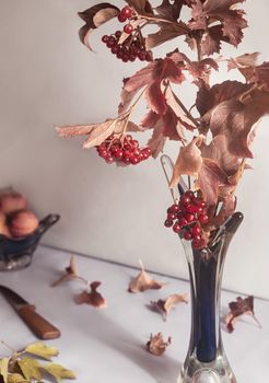 On the table in a glass vase is a branch of viburnum with ripe berries and red autumn leaves. Front view, close-up.