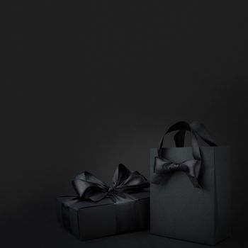 Concept sales, shopping, black friday. Paper shopping or gift bag box with silk ribbon bow on black background with copy space