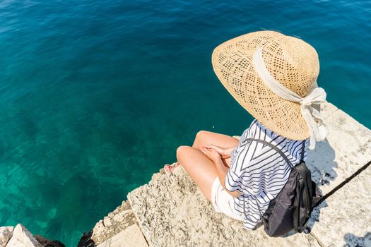 Woman traveler wearing straw summer hat and backpack, standing at edge of the rocky cliff looking at big blue sea and islands in on the horizon.