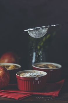 Sifting powdered sugar by sieve over apple pie in ceramic baking molds ramekin on dark wooden table. Close up, shallow depth of the field 