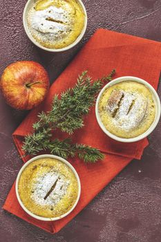 Three apple pies in ceramic baking molds ramekin with napkin, apple, spruce branch, cinnamon on dark red concrete table. Top view, flat lay. Christmas and New Year food background.