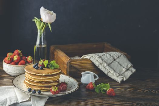 Pancake with strawberry, blueberry and mint in ceramic dish, syrup from small ceramic jar and flowers on a dark wooden table