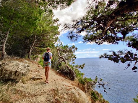 Young active feamle tourist wearing small backpack walking on coastal path among pine trees looking for remote cove to swim alone in peace on seaside in Croatia. Travel and adventure concept.