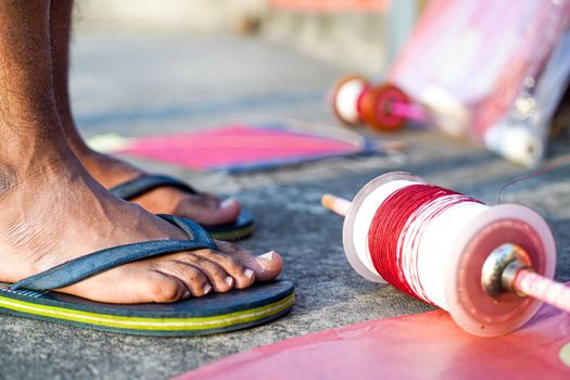 Young man with rubber slippers chappal with a charkhi fikri spool with dor thread with glass thread and kites placed nearby on the indian festival of makar sankranti uttarayana . This festival is known for its colorful kites of paper and wood used for kite fighting with a glass covered thread to cut an opponents kite