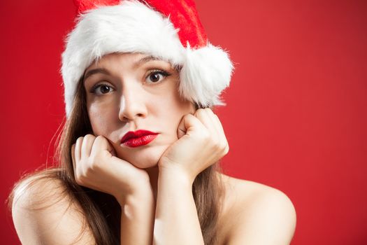 Worried depressed attractive caucasian brunette woman dressed as Mrs Santa Claus,wearing red white hat,chin on hands,isolated on red background,copy space,New Year 2021 COVID-19 Coronavirus pandemic