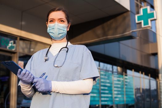 Young female doctor standing in front of healthcare facility, wearing protective face mask and PPE equipment, holding medical patient clipboard, COVID-19 pandemic crisis