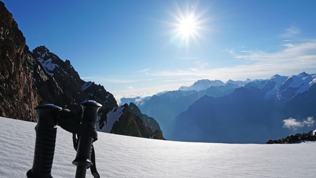 Trekking sticks on the background of snow peaks. The sun is shining brightly, white clouds and snow cliffs in the distance. Blue clear sky. Climbers are going to the top. Alpine camp in the mountains.