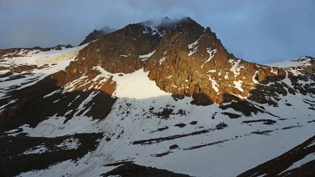 View of the rocky slopes covered with snow. The sun's rays and clouds. Mountaineering camp in the mountains. There is a nunatak. Overnight in the mountains. Sunset in the cold mountains.